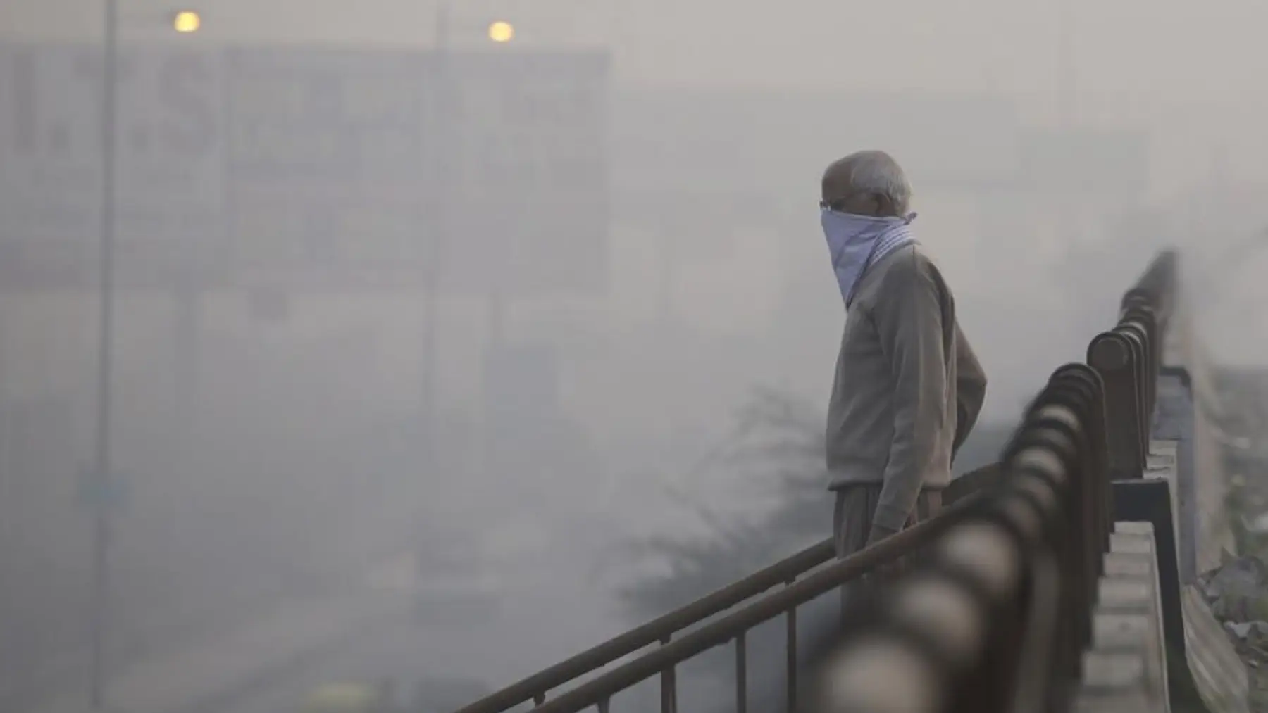 A man with his face covered walks along a railing in thick grey smog